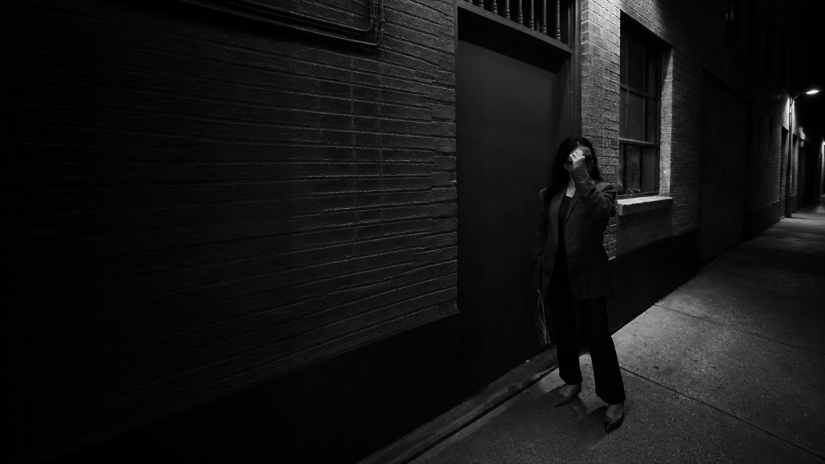 A woman in black tailoring stands in a dark brick alley, framed by deep architectural shadows.