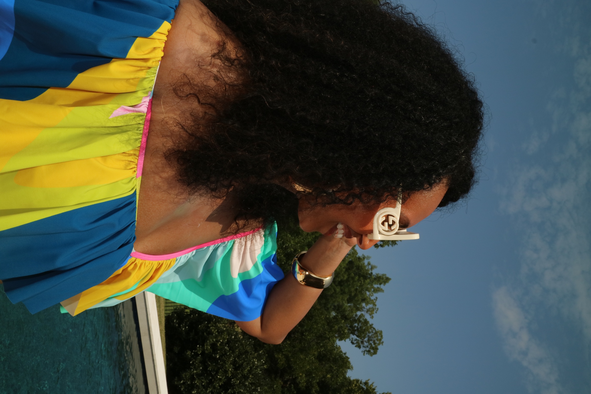 Poolside portrait of Lela wearing white sunglasses and a vivid multicolor dress beneath a blue sky.