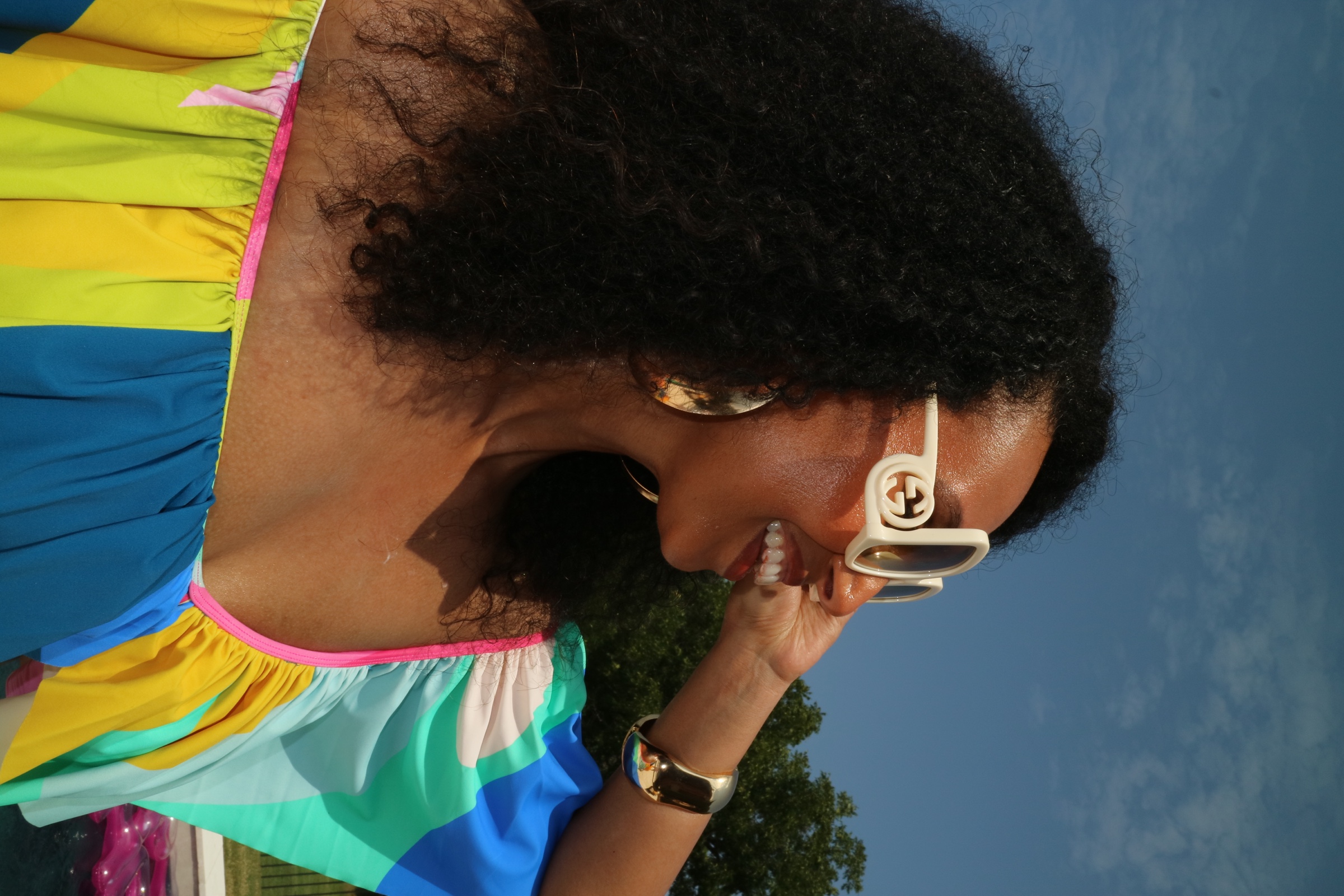 Close poolside portrait of Lela smiling in white sunglasses and a vivid multicolor dress beneath a blue sky.