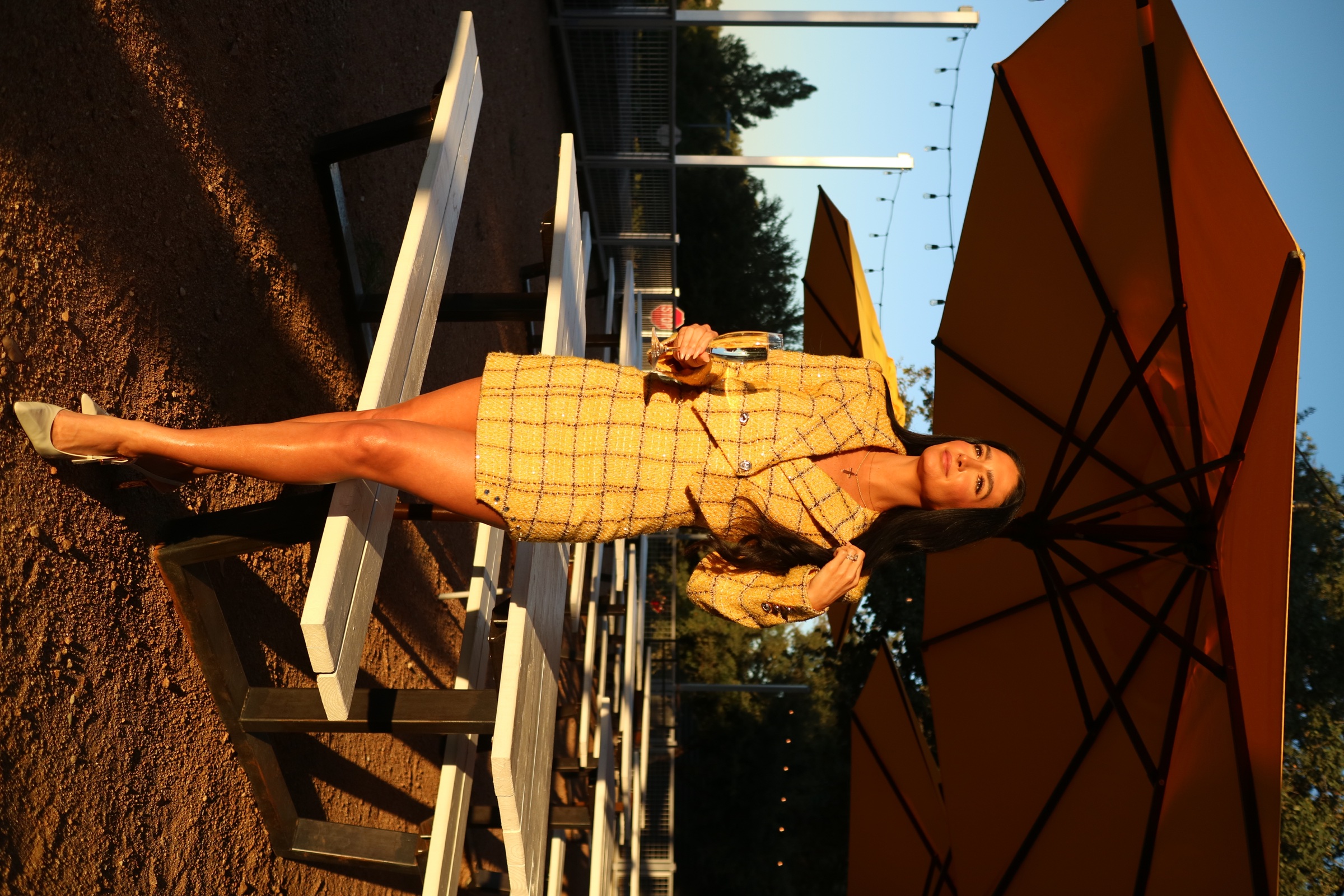 Golden-hour fashion portrait of a woman in a yellow checked suit holding a champagne glass beneath patio umbrellas.