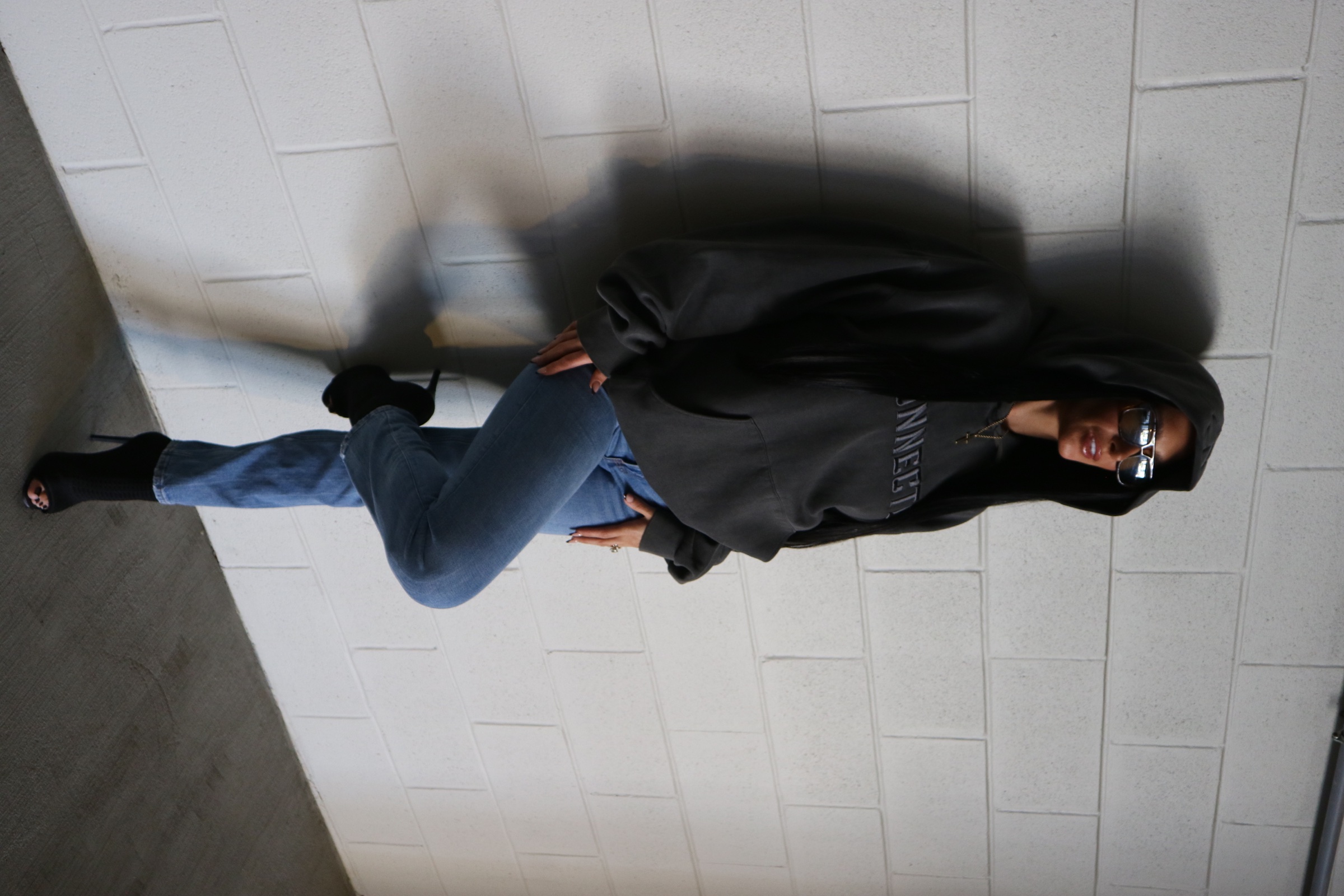 Fashion portrait of a woman in a charcoal hoodie and jeans leaning against a concrete block wall.